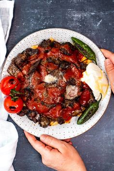 two hands holding a plate of food with tomatoes and other foods on the table next to it