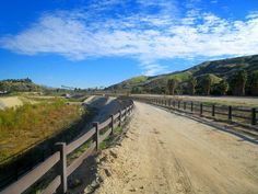 a dirt road with a wooden fence next to it and hills in the background on a sunny day