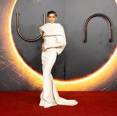 a woman in white dress posing for the camera on a red carpet with an orange circle behind her