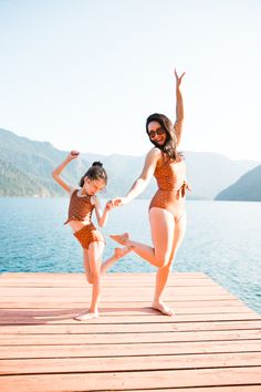 two women in bathing suits are standing on a dock near the water and stretching their arms