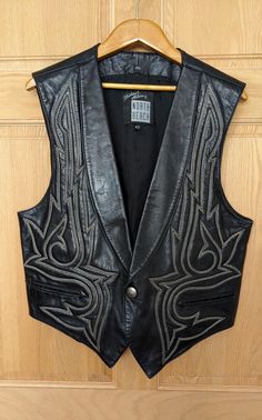 Wonderful black North Beach Leather Vest by Michael Hoban with Western Cowboy style stitching, snap and back belt buckle.  The leather is very soft and pliable, and there's slight padding behind the stitching in front.  There are 2 pockets on the outside of the vest and 2 more on the inside lining, as shown in the photos.  The only flaw I can find is a slight rough spot and 2 tiny pin dots on the right lapel.  They look more prominent in the photos than in real life.  Otherwise, the vest is in excellent condition.  A size 42, the measurements are approx:   16 1/2' across the shoulders, 42" across the chest   40" across the bottom    25" long in front to the vest tips   22" long in the mid back See my entire shop collection here https://www.etsy.com/shop/RobinClaytonVintage Western Denim Vest, Men’s Vests, Gothic Cowboy Fashion, Goth Cowboy Men, Unique Outfits Men, Black Cowboy Outfit, 70s Rock Style, Edgy Western Style, Cowboy Outfit Men