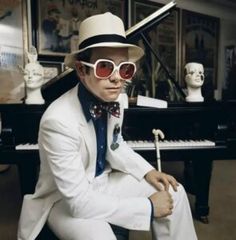 a man in a white suit and hat sitting next to a piano wearing red sunglasses