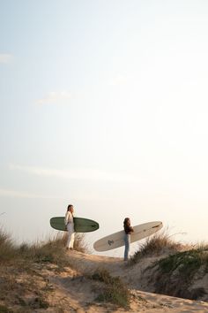 two people carrying surfboards on top of a sandy hill at the ocean's edge