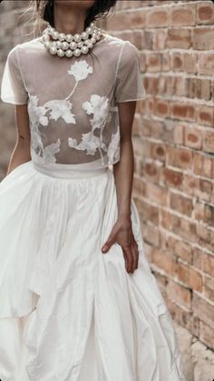 a woman in a white dress standing next to a brick wall and wearing a necklace