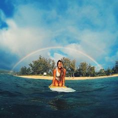 a woman is sitting on a surfboard in the water with a rainbow behind her