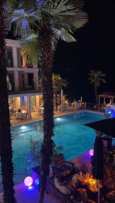 an outdoor swimming pool at night with palm trees in the foreground and lights on