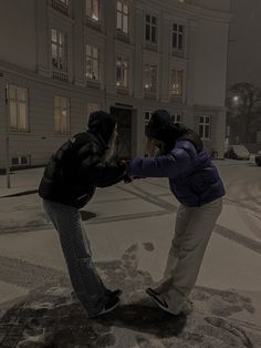 two people standing in the snow touching each other's hands with buildings in the background