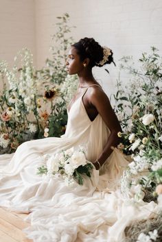 a woman in a white dress is sitting on a bed with flowers and greenery