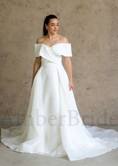 a woman wearing a white wedding dress standing in front of a concrete wall with her hands on her hips