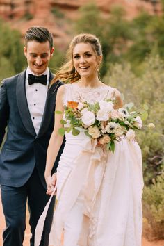 a bride and groom holding hands while walking through the desert in their tuxedos