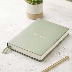 a green notebook sitting on top of a table next to a pen and potted plant