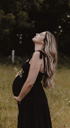 a pregnant woman in a black dress standing in tall grass looking up at the sky