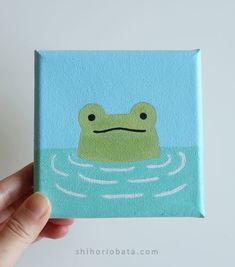 a hand holding up a small square painting with a frog on it's face