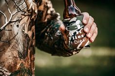 a man holding a beer bottle in his right hand and wearing camouflage clothing with trees on it