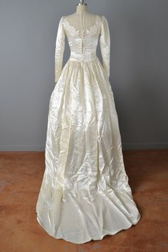 "This stunning gown from the 1940s is an ivory satin and features a long train, drop waist with gathers that give the hip and the skirt fullness. The bodice consists of a fine net illusion neckline, which has been appliquéd with large trapunto flowers in the front and back. Covered satin buttons up the back, tapered long sleeves with covered buttons, and gentle puffed shoulders. Measurements → Size: Small Bust: 34\" Waist: 26\" Hips: Free Length (front): 58\" Length (tip of the train): 89\" Cond Full Length Bias Cut Gown For Wedding, Vintage Wedding Gown With Bias Cut, Vintage Satin Wedding Gown, Vintage Cream Satin Gown, Vintage White Wedding Dress With Fitted Bodice, Cream Vintage Gown For Debutante Ball, Vintage Ball Gown With Boned Bodice, Cream Vintage Wedding Dress With Fitted Bodice, Vintage Gown With Fitted Bodice For Wedding Night