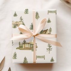 a present wrapped in white wrapping paper with trees and houses on it, tied with a pink ribbon