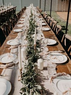 a long wooden table with white plates and greenery on it, surrounded by candles