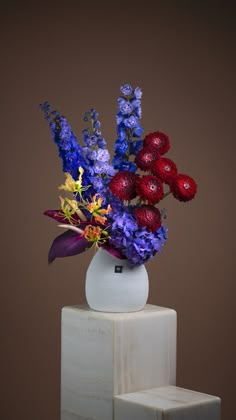 a white vase filled with colorful flowers sitting on top of a wooden block in front of a brown wall