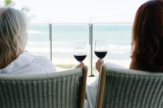 two women sitting on chairs with wine glasses in their hands looking out at the ocean