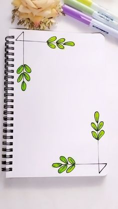 a spiral notebook with green leaves on it next to two markers and a flower arrangement