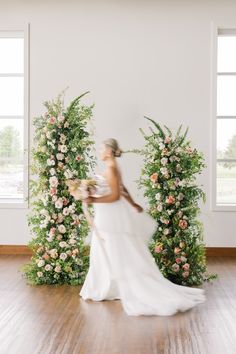 a woman in a white dress standing next to flowers and greenery on a wooden floor