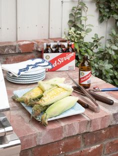 corn on the cob and beer are sitting on a brick counter top next to an outdoor bbq