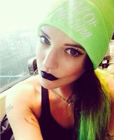 a woman with long green hair wearing a beanie and black nose ring in front of a window