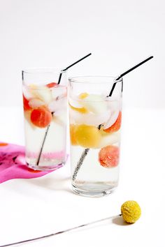 two glasses filled with ice and fruit on top of a white table next to a pink spoon