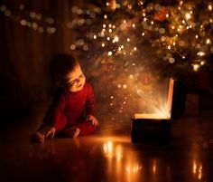 a baby sitting on the floor next to a christmas tree looking at an open box