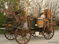 an old fashioned wagon with many items on it's back and wheels, sitting in front of some trees