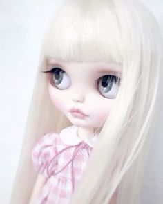 a white doll with long blonde hair and big blue eyes wearing a pink dress is standing in front of a white wall