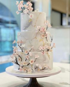 a three tiered white cake with pink flowers on the side and gold accents, sitting on a marble stand