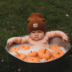 a baby in a bucket filled with pumpkins on top of green grass and wearing a brown hat