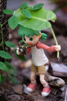 a small doll holding a camera next to a tree with leaves on it's head