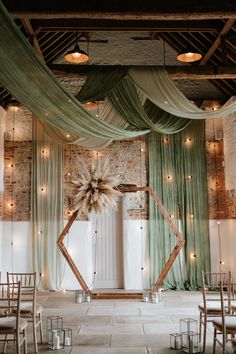 This shows a ceremony area at Curds Hall Barn, Norfolk. The focus is on a hexagon backdrop at the end of the aisle with a dried flower arrangement on the top left corner. At the base are some black box lanterns with candles. Behind and above the hexagon are green ceiling drapes paired with festoon wall lighting. Rustic Wedding Set Up Receptions, Sage Wedding Backdrop, Wedding Backdrop Drapes, Barn Draping Wedding, Modern Barn Wedding Decor, Green Wedding Backdrop, Unique Wedding Backdrop, Hexagon Backdrop, Activation Booth