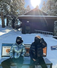 two people sitting on a bench in the snow near a small building with a sign