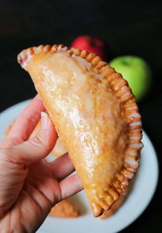 a hand holding a pastry on top of a white plate next to an apple slice