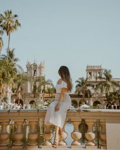 a woman in white dress standing on railing next to water and palm trees with buildings in the background