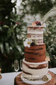 a three tiered wedding cake with flowers and greenery sits on a tree stump