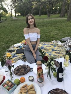 a woman is sitting on a blanket with food and drinks in front of her at a picnic