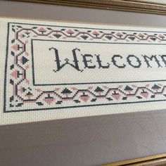 a cross - stitch welcome sign hanging on a wall in front of a framed photo