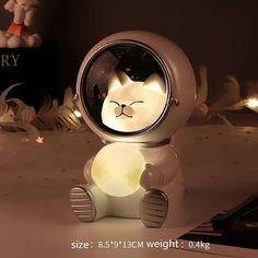 a white cat sitting on top of a table next to a lamp that is shaped like an astronaut's helmet