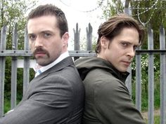 two men are sitting on a bench in front of an iron fence and looking at the camera