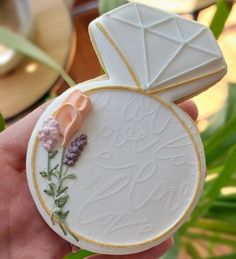 a hand holding a white and gold decorated cookie with flowers on the inside, in front of a plant