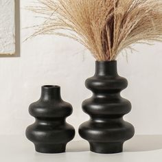 two black vases with grass in them on a table