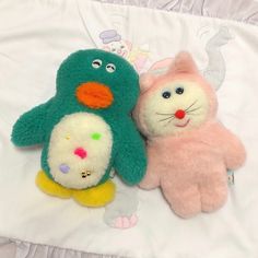 two stuffed animals laying on top of a white bed sheet with pink and green colors