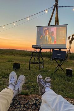 two people are sitting in the grass watching tv with their feet propped up on a blanket