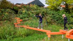 two children playing on an orange bench in the grass with trees and bushes behind them