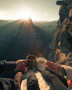 two people sitting on the edge of a cliff with their feet up holding coffee cups