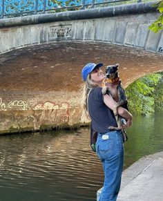 a woman kissing a dog on the side of a river under a bridge with graffiti